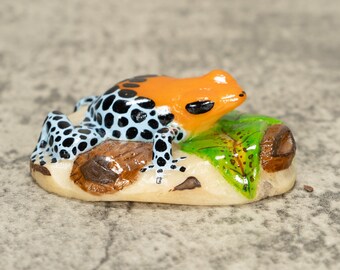 May include: A small, hand-painted ceramic figurine of a poison dart frog sitting on a leaf. The frog is orange, black, and blue with white spots. The leaf is green with brown veins.