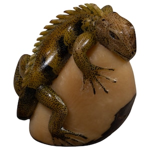 Puede incluir: Una figura tallada de una iguana con cuerpo de color verde oliva moteado de negro, que presenta una cresta dorsal prominente. El lagarto está posado sobre una base de piedra lisa y redondeada de color crema con una mancha marrón oscura.