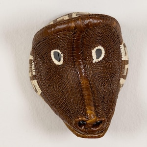 May include: A brown woven mask with white accents around the eyes and mouth. The mask is shaped like an animal face with a long snout.
