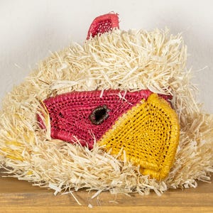 Puede incluir: Una máscara de pájaro artesanal hecha de paja y rafia tejidas. La máscara presenta un pico rojo y amarillo, con un ojo oscuro. La máscara está rodeada de un material esponjoso similar a la paja.