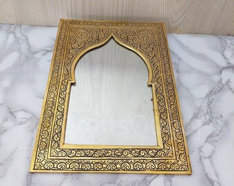 May include: A gold-coloured metal mirror with an ornate, black-etched frame. The frame features a traditional Moroccan arch design with intricate floral patterns.