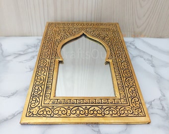 May include: A gold-coloured metal mirror with an ornate black etched design around the frame. The mirror has an arched opening in the centre.