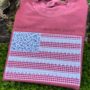 May include: A dusty rose t-shirt featuring a rectangular graphic of an American flag made from fabric patches. The flag has a blue floral canton and red and white gingham stripes accented with white lace trim. The text MNM Embroidery and Design is visible.