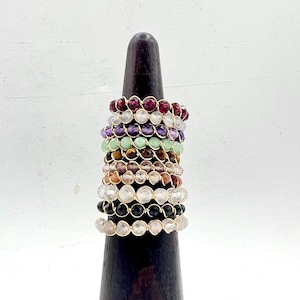 May include: A stack of 12 colorful beaded rings on a wooden ring display stand. The rings are made with different colored beads, including red, purple, green, brown, and black.