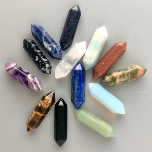 May include: A collection of 12 crystal wands in various colors, including amethyst, blue, green, black, brown, and white. The wands are all pointed at one end and have a smooth, polished finish.