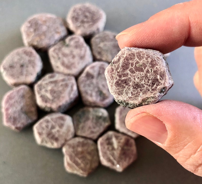 K&ouml;nnte beinhalten: Eine Sammlung von rohen rosa Rubin-Steinen, wobei ein Stein in einer Hand gehalten wird. Die Steine sind facettiert und haben ein nat&uuml;rliches, rohes Aussehen.