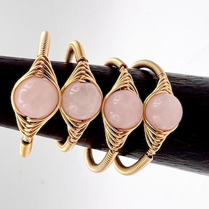 May include: Four gold wire wrapped rings with pink rose quartz stones. Each ring features a delicate wire design and a smooth, round stone.