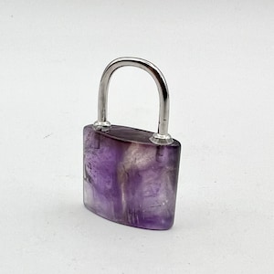 May include: A small, purple amethyst padlock with a silver metal loop. The padlock is made of polished amethyst and has a smooth, rounded shape.