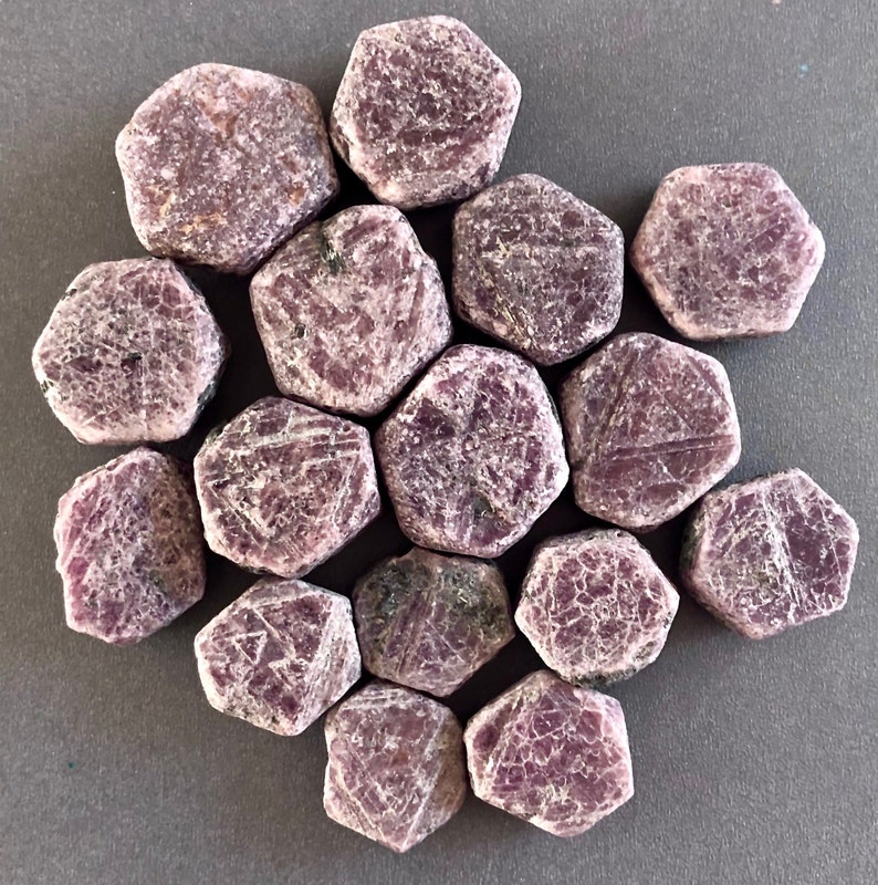 K&ouml;nnte beinhalten: Eine Sammlung von 14 facettierten, sechseckigen, rosafarbenen Rubinen. Die Steine sind in einem Kreis auf einer grauen Oberfl&auml;che angeordnet.