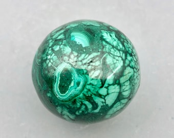 May include: A polished spherical stone featuring a complex pattern of deep forest green and lighter mint green swirls. The surface displays intricate concentric banding and organic web-like veins characteristic of malachite, with a smooth, reflective finish.