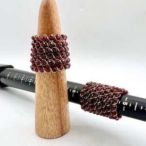 May include: Two red beaded rings with gold wire. The rings are on a wooden ring mandrel and a black ruler.