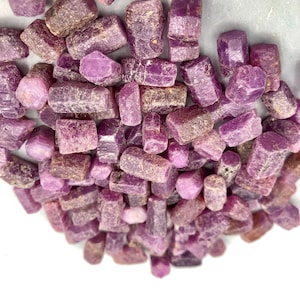 May include: A collection of small, rough, purple ruby crystals. The crystals are various shapes and sizes, and have a natural, unpolished look.