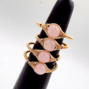 May include: Four gold wire wrapped rings with pink rose quartz stones. The rings are stacked on a wooden ring holder.