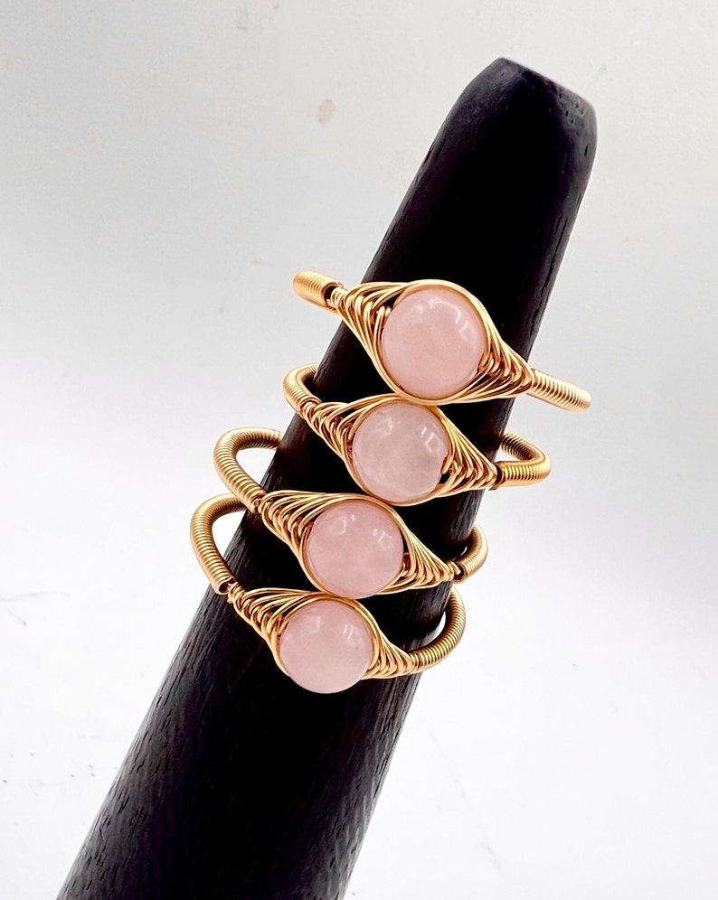 May include: Four gold wire wrapped rings with pink rose quartz stones. The rings are stacked on a wooden ring holder.