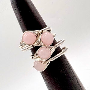 May include: Three silver wire wrapped rings with pink rose quartz stones. The rings are stacked on a wooden ring display stand.