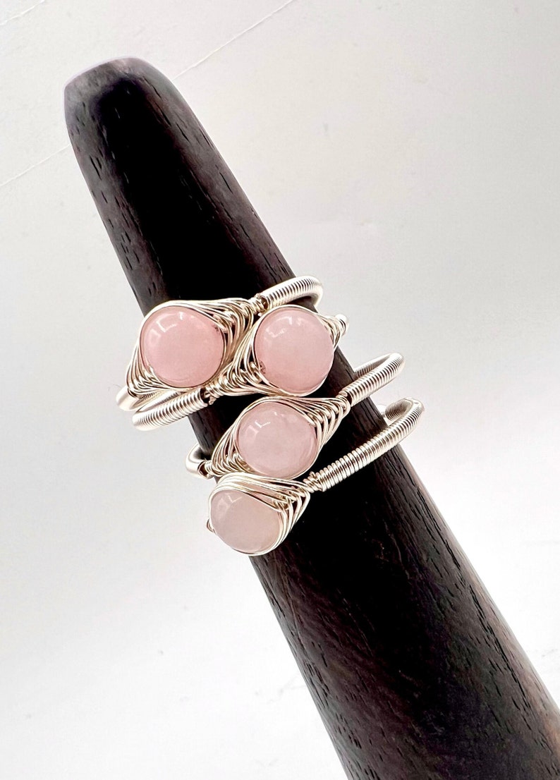 May include: Three silver wire wrapped rings with pink rose quartz stones. The rings are stacked on a wooden ring display stand.