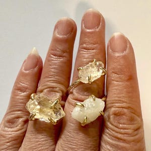 May include: Three gold rings with clear quartz stones. The rings are worn on the fingers of a hand.