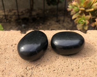 May include: Two smooth, black obsidian stones, polished to a high shine, are displayed on a light brown surface. The stones are oval-shaped and have a natural, organic feel.