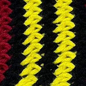 May include: Close-up of a colorful crochet pattern. The design features alternating rows of blue, red, gray, black, and yellow yarn, creating a textured, zig-zag effect. The vibrant colors and detailed stitching suggest a handmade textile.