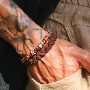 Op de afbeelding: Een set van twee armbanden bestaande uit een gevlochten donkerbruin leren koord en een kralenarmband met afwisselende bruine houten kralen en kleine zilverkleurige metalen tussenstukjes, samen gedragen om de pols.