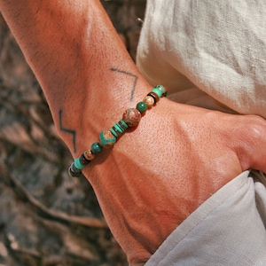May include: A green and brown beaded bracelet with a gold accent. The bracelet is worn on a person's wrist.