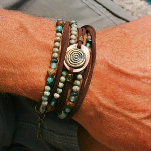 Op de afbeelding: Een meerlaags armband met bruin lederen koord, turquoise en groene kralen en een koperkleurige ronde bedel met een spiraalvormig ontwerp.