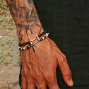 Puede incluir: Dos pulseras de cuentas con una mezcla de cuentas negras, marrones y color crema. Las pulseras se llevan en una muñeca con un tatuaje. Las pulseras tienen detalles metálicos plateados.