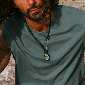 May include: A sleeveless, olive green shirt. A black leather necklace with a pendant featuring a colorful, patterned design hangs around the neck. The person has tattoos on their arm.