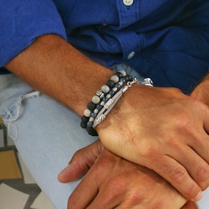 Op de afbeelding: Een mannenpols met drie armbanden. De armbanden zijn gemaakt van donkerblauwe en grijze stenen, en een heeft een zilveren bedel. De man draagt ook een zilveren ring aan zijn vinger.