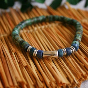 Op de afbeelding: Een armband met turquoise en blauwgroene kralen, geaccentueerd met zilverkleurige metalen kralen en een rechthoekige sluiting. De armband wordt getoond op een getextureerd, lichtbruin oppervlak.
