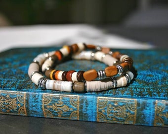 Pode incluir: Duas pulseiras sobrepostas com contas marrons, brancas e bege. As pulseiras estão sobre um livro azul com detalhes dourados.