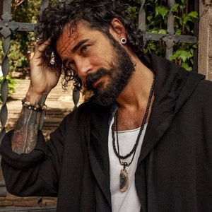 May include: A man with curly dark hair and a beard is wearing a black hooded jacket. He has a black beaded necklace with a large, natural stone pendant. He is also wearing a bracelet on his left wrist.