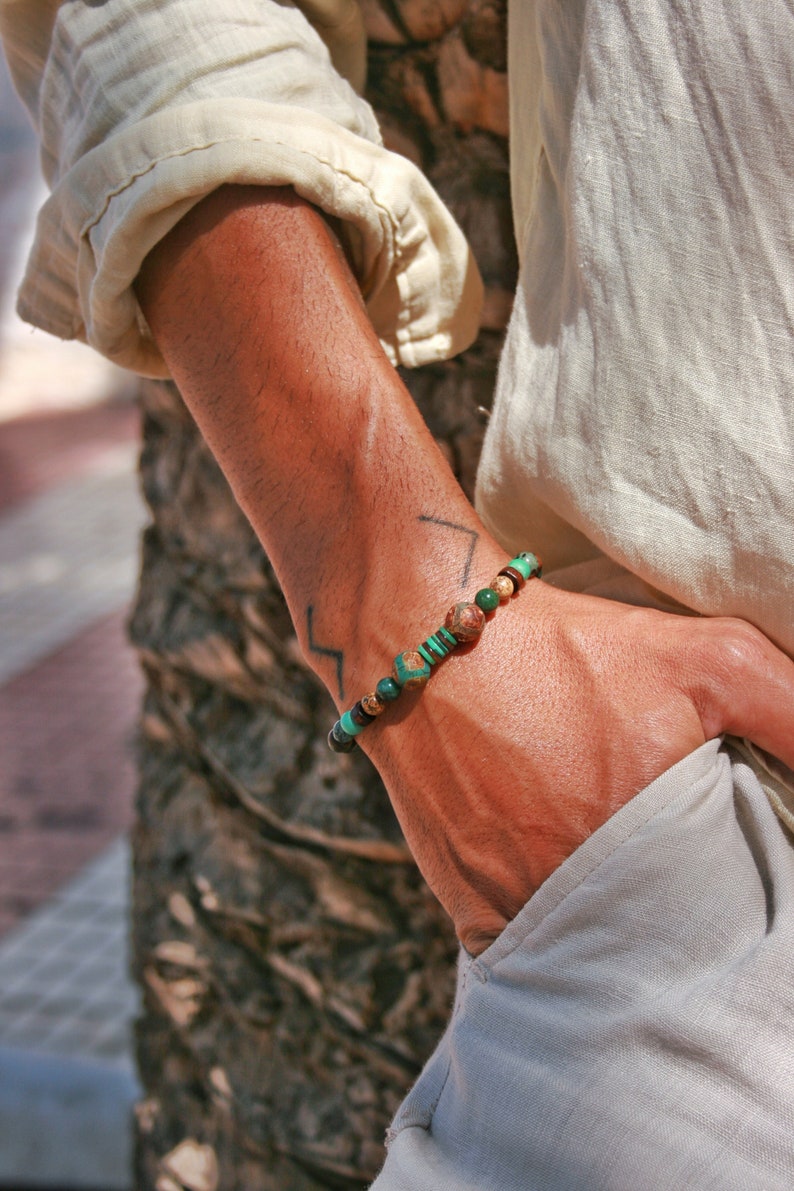 May include: A green and brown beaded bracelet with a gold accent. The bracelet is worn on a person's wrist.