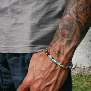 Könnte beinhalten: Ein Armband mit türkisfarbenen, braunen und silbernen Perlen. Das Armband wird am Handgelenk getragen, mit einem grauen Oberteil und einer marineblauen Hose im Hintergrund. Der Arm der Person ist tätowiert.