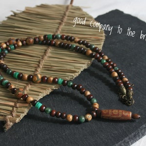 May include: A brown and green beaded necklace with a wooden pendant. The pendant has a carved design and is attached to the necklace with a metal clasp.