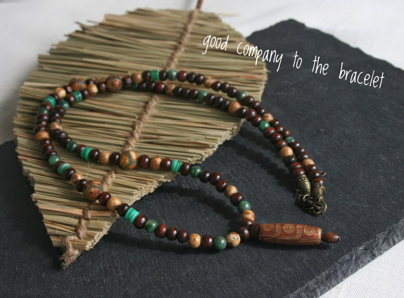 May include: A brown and green beaded necklace with a wooden pendant. The pendant has a carved design and is attached to the necklace with a metal clasp.