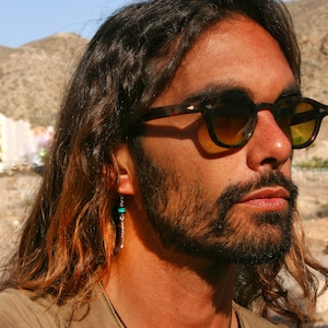 Può includere: Un uomo con i capelli lunghi castani e la barba indossa occhiali da sole in tartaruga marrone. Ha un orecchino di pietra verde nell'orecchio sinistro.
