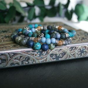 Puede incluir: Un conjunto de cuatro pulseras de cuentas en tonos azules y verdes. Las pulseras están hechas de varios materiales, incluyendo piedra, madera y metal. Las pulseras están dispuestas sobre una superficie decorativa.