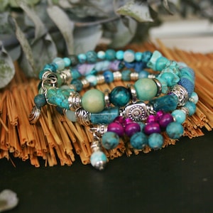 May include: A set of four turquoise and purple beaded bracelets with silver accents. The bracelets are stacked on a woven straw mat.