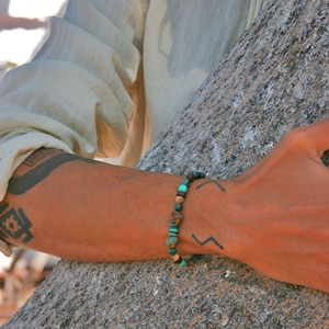 May include: A close-up of a person's wrist wearing a beaded bracelet with brown, green, and turquoise beads. The bracelet is on a person's left wrist and the person is wearing a white shirt.