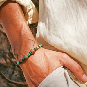 May include: A close-up of a person's wrist wearing a beaded bracelet with green, brown, and black stones. The bracelet is made of natural materials and has a simple, elegant design.