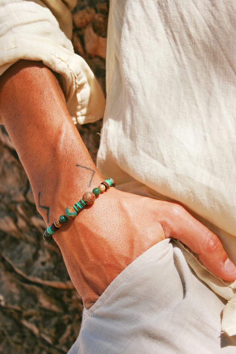 May include: A close-up of a person's wrist wearing a beaded bracelet with green, brown, and black stones. The bracelet is made of natural materials and has a simple, elegant design.