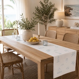 May include: A white rectangular table runner featuring a repeating blue line-art pattern of sailboats, lighthouses, fish, and botanical sprigs, laid flat across a wooden dining table with a textured surface and finished edges.