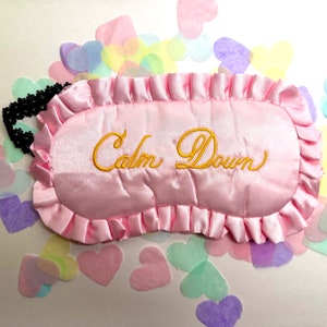 May include: Pink satin sleep mask with a ruffled edge and the words "Calm Down" embroidered in gold thread.