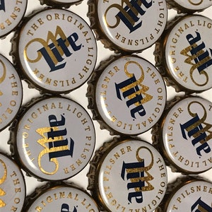 May include: Close-up of multiple gold and white bottle caps with the text "The Original Light Pilsner Lite" printed on them.
