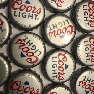 May include: A collection of silver bottle caps with the Coors Light logo. The caps are arranged in a random pattern and feature the text "Cold as the Rockies" and a mountain peak graphic.