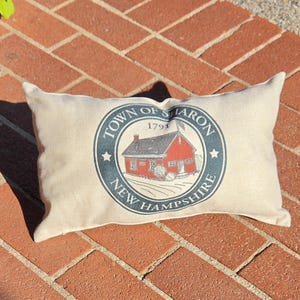 May include: A rectangular beige throw pillow featuring a circular graphic in the center. The graphic depicts a red building with the text "TOWN OF SHARON 1791 NEW HAMPSHIRE" printed in dark blue lettering around the perimeter of the circle.