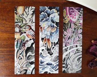 May include: Three watercolor bookmarks featuring different designs. The first bookmark features a bird perched on a branch with lemons and leaves. The second bookmark features a blue flower with a wave pattern. The third bookmark features a sword with a pink flower and leaves.