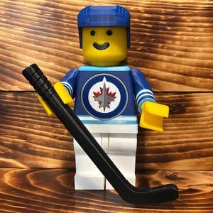 May include: A Lego minifigure dressed as a hockey player wearing a blue and white jersey with the Winnipeg Jets logo. The figure is holding a black hockey stick.