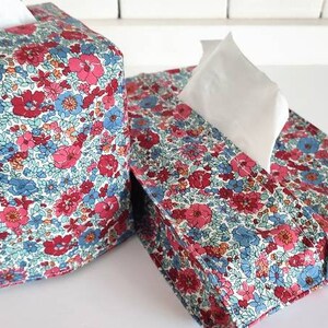 May include: Two floral patterned tissue box covers. The covers are made of fabric with a white background and a floral pattern in shades of pink, blue, and green. The covers are square and have a slot for tissues to be pulled out.